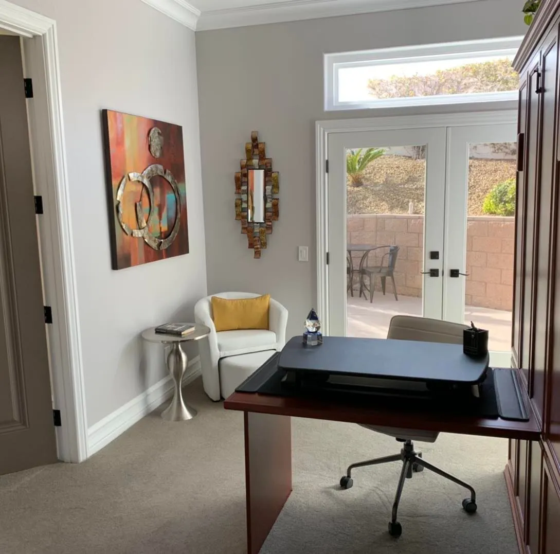 Elegant home office featuring a dark wood desk, modern art on the walls, and a comfortable white armchair, with sunlight streaming in through French doors leading to a patio